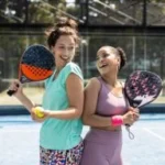 Two women wearing athletic attire stand back-to-back on a paddle tennis court, smiling and holding paddles. One holds a yellow ball. The court is enclosed with a fence.