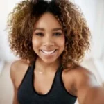 A person with curly hair wearing a sleeveless black top smiles while taking a selfie indoors.