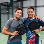 Two men in sports attire holding padel rackets, standing side by side on a padel court, smiling at the camera.